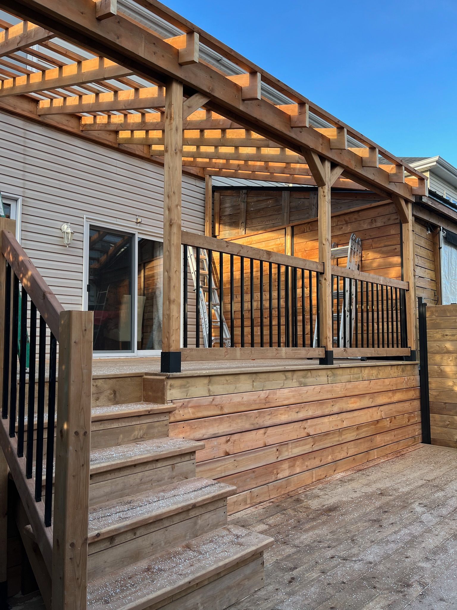 Terrasse en bois avec pergola, escalier et rambarde noire, adossée à une maison sous un ciel bleu azur.