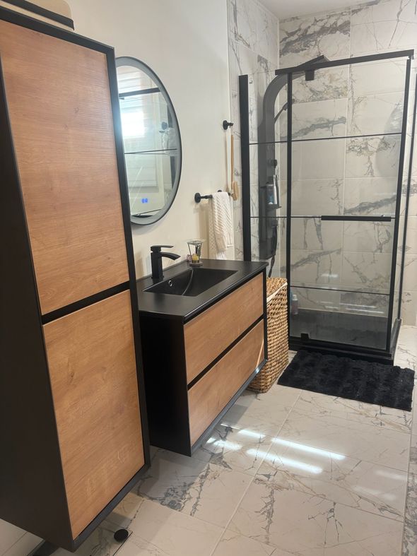 Salle de bains moderne avec douche noire, meuble-lavabo en bois, miroir ovale et armoire haute.