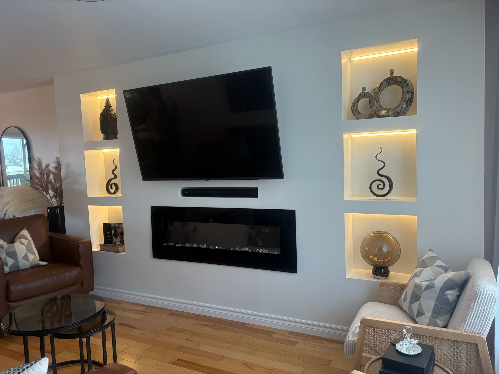 Salon avec cheminée intégrée, télévision et niches murales éclairées présentant des objets décoratifs ; parquet, murs clairs et deux fauteuils.