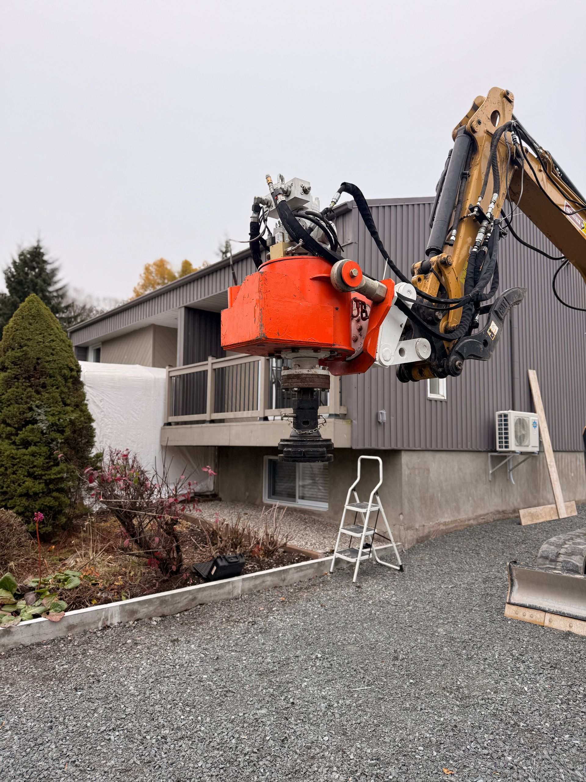 Une pelleteuse équipée d'un accessoire orange en train d'enlever quelque chose. Bâtiment gris, allée de gravier, ciel couvert.