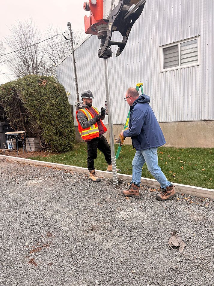 Deux ouvriers installent un poteau à l'aide d'engins lourds sur un sol gravillonné près d'un bâtiment. L'un semble diriger, l'autre l'assiste.