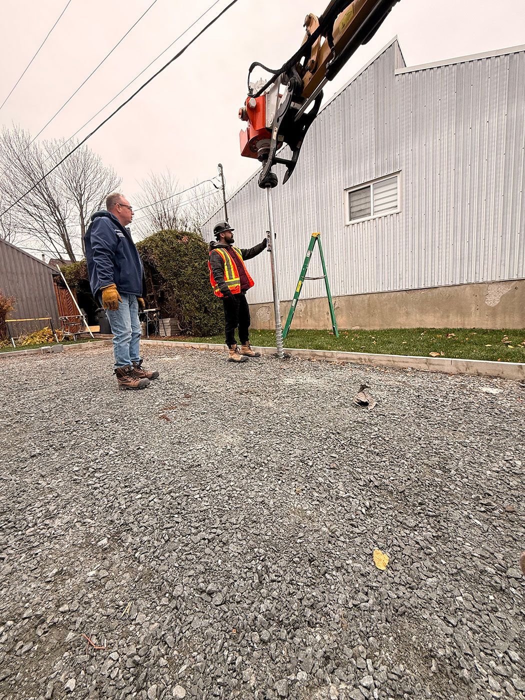 Deux ouvriers manipulent des machines près d'un bâtiment. L'un dirige, l'autre observe. Une grue est installée au-dessus.