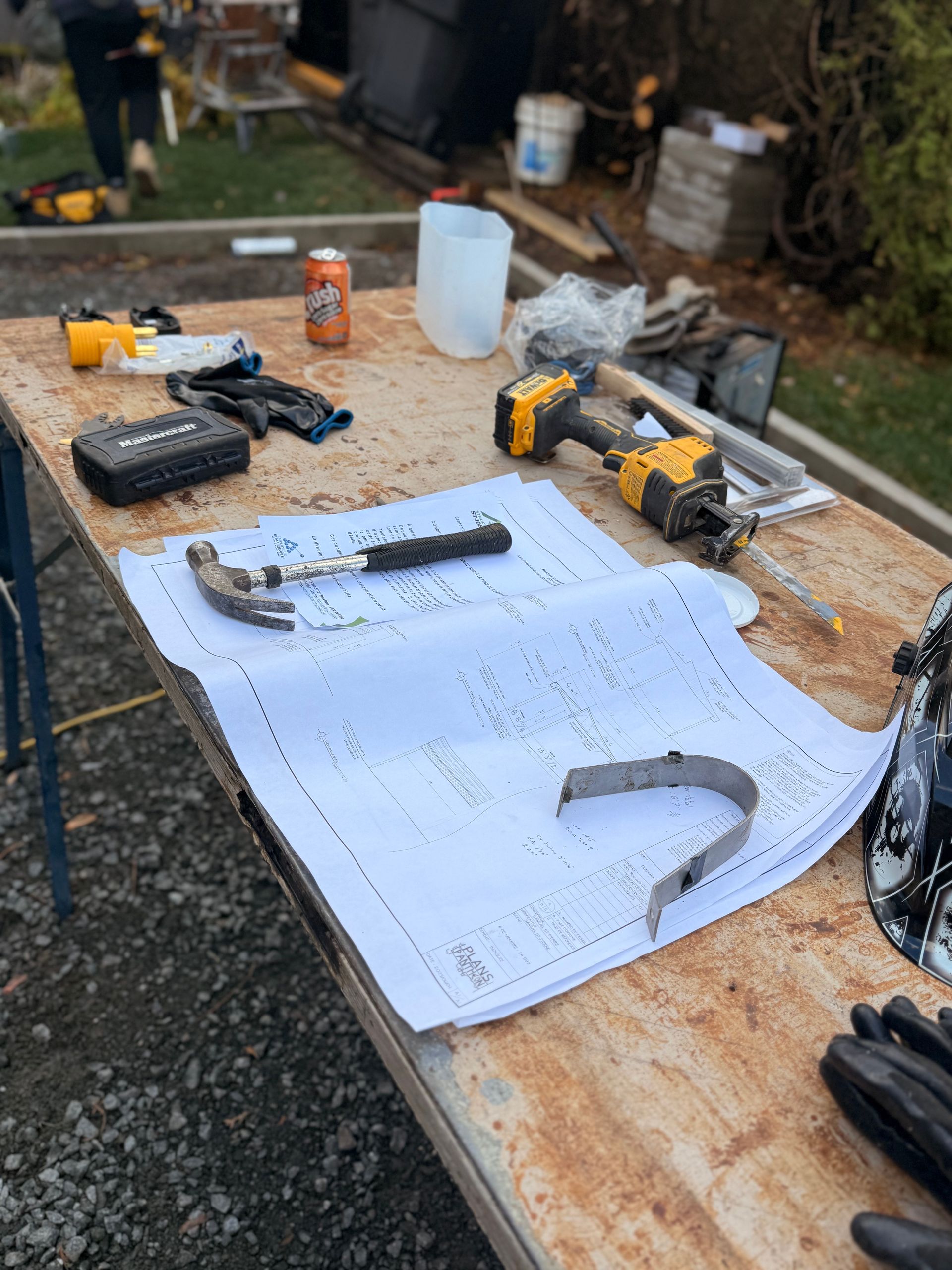 Table de travail avec plans, outils, canette de soda et pièce de métal, à l'extérieur.