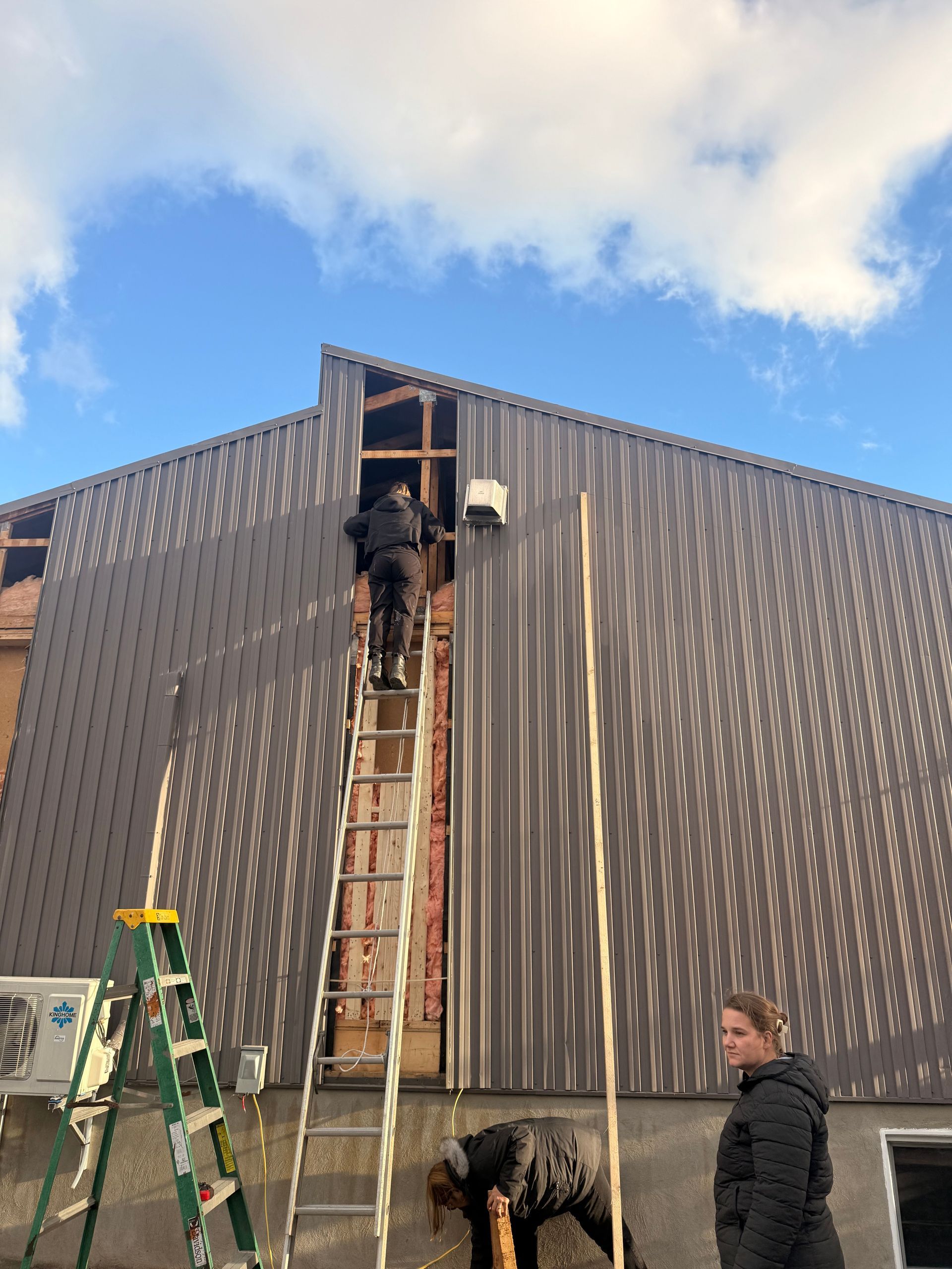 Une personne sur une échelle travaille sur le côté d'un bâtiment, bardage brun, ciel bleu nuageux.