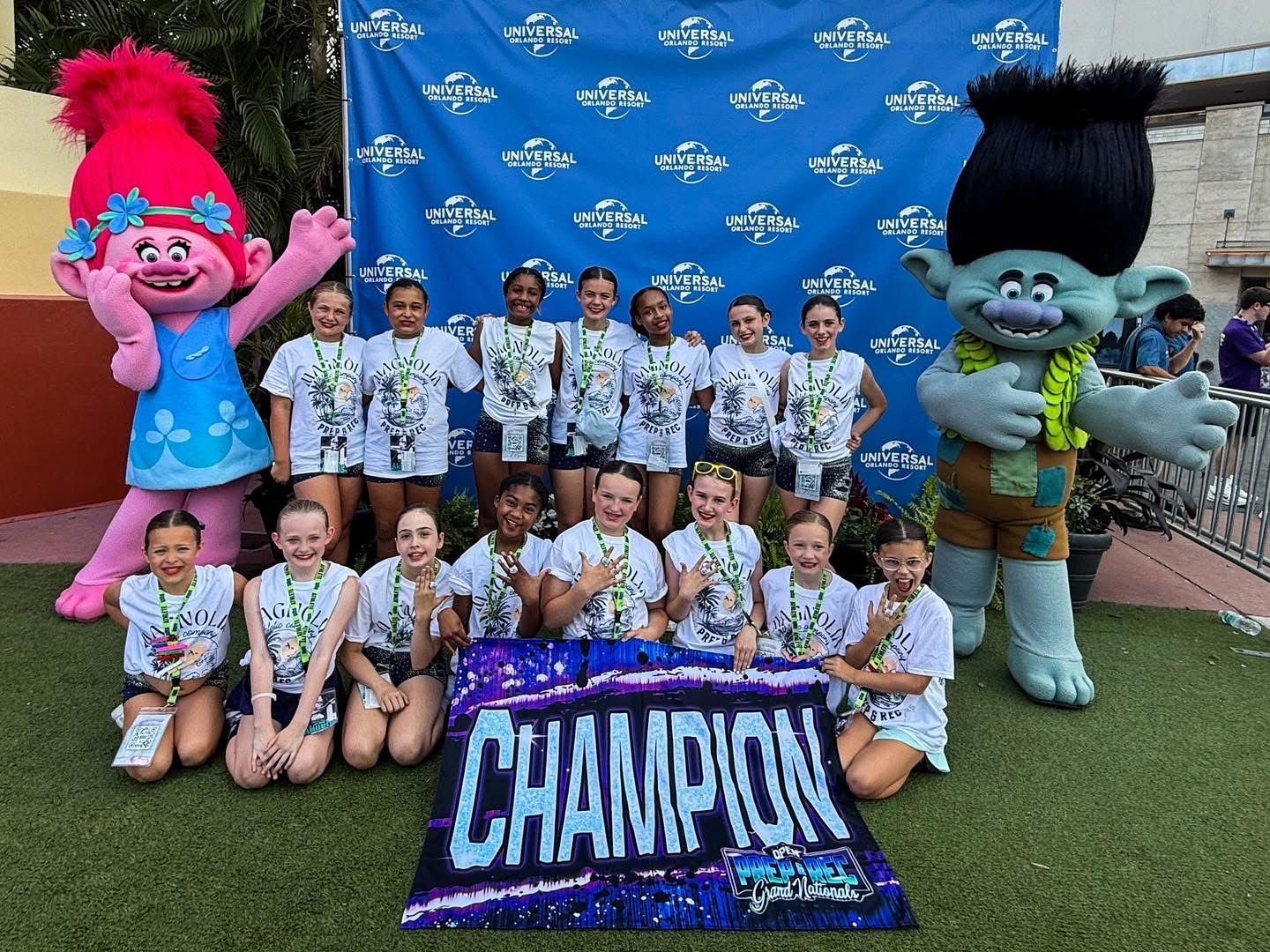 Dance team poses with trophies and mascots. Blue backdrop, green turf, sunny.