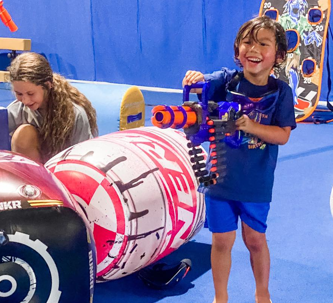 Boy smiles, holding Nerf gun, aiming at inflatable target. Girl looks on. Blue floor.