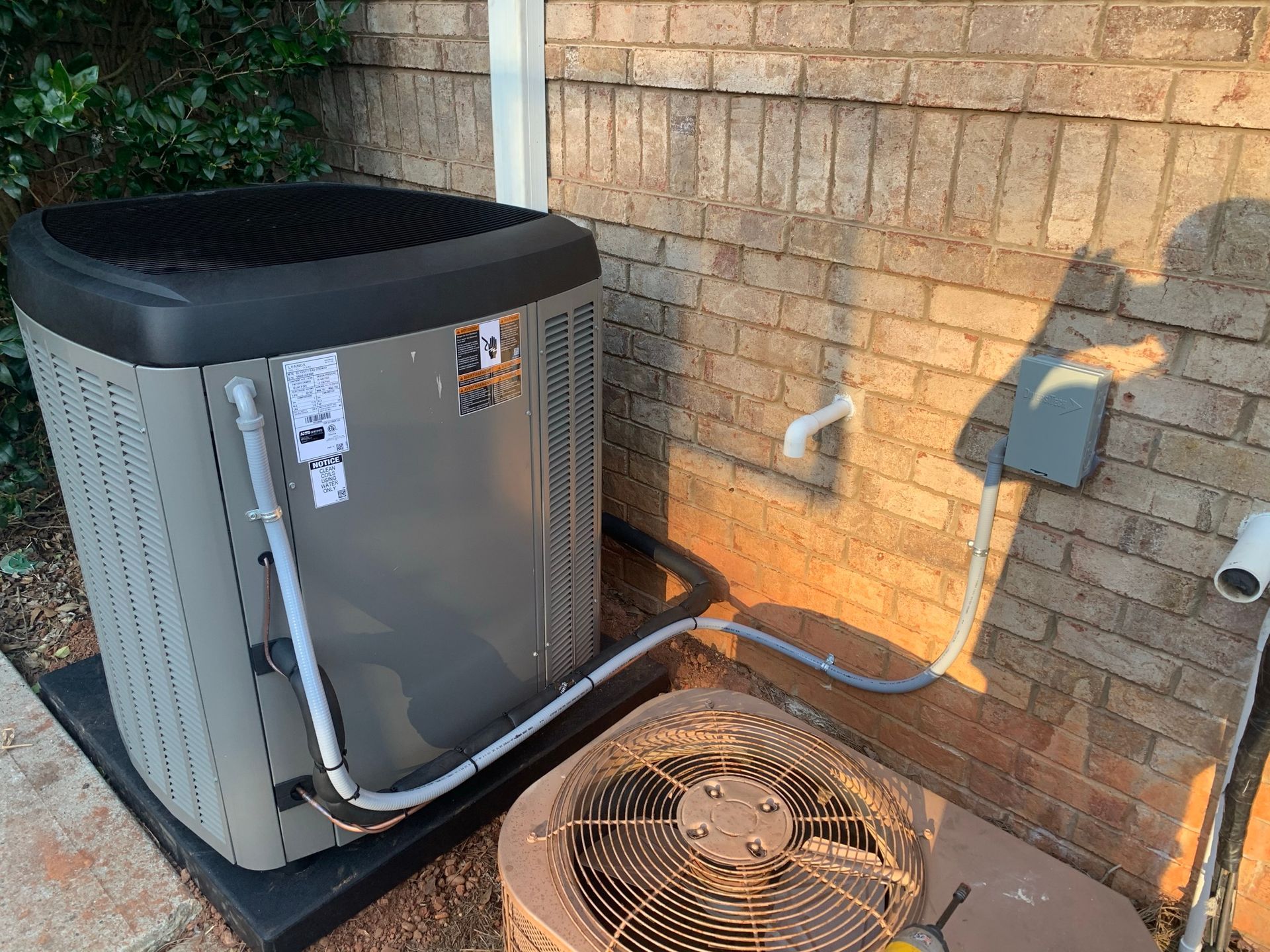 An outdoor air conditioning unit next to a brick wall. A smaller fan is below it, with electrical and piping connecting the units.