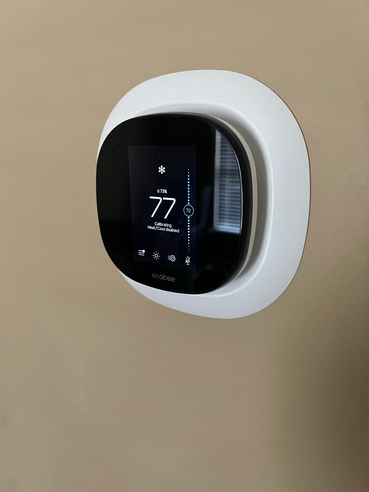 A smart thermostat, displaying 77 degrees, mounted on a beige wall, surrounded by a white plate.