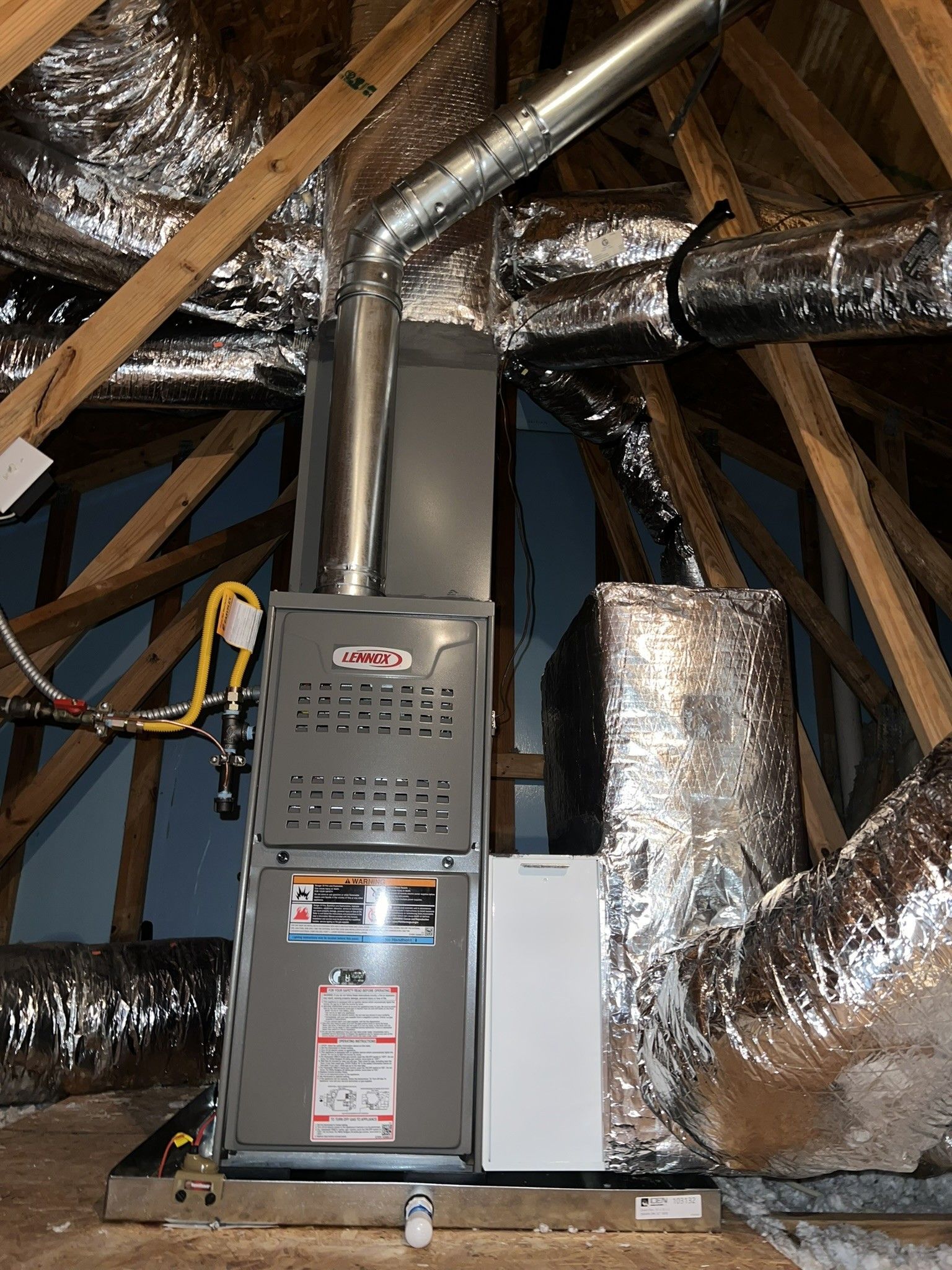 Furnace unit installed in an attic with ductwork, vent pipe, and a white filter.