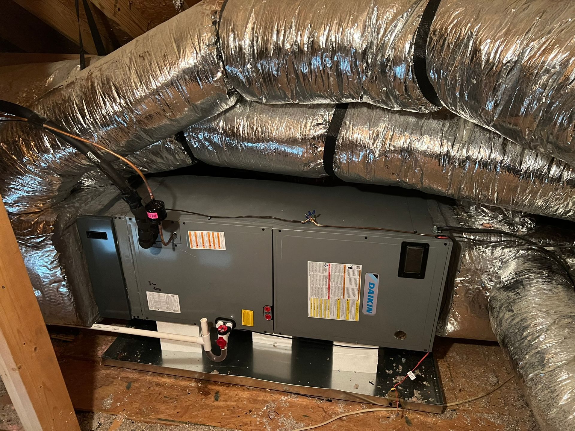 HVAC unit in an attic with insulated ductwork. Gray unit on metal stand, surrounded by silver tubes.