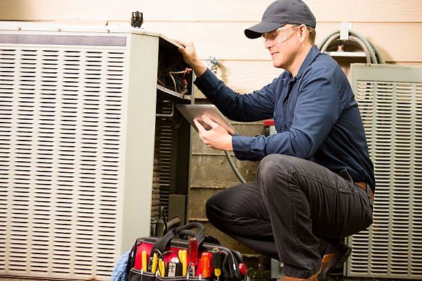 A technician in a cap and work shirt inspects the open panel of an outdoor air conditioning unit near a tool bag.