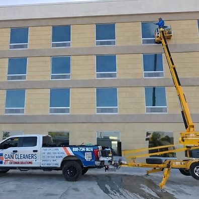 Worker in a yellow lift cleaning windows on a beige building beside a blue window-cleaning van.