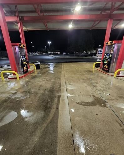 Rainy gas station car wash bay at night, wet concrete floor reflecting overhead lights.