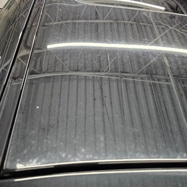 Close-up view of a dark car roof with a sunroof, reflecting bright overhead warehouse lighting and metal ceiling beams.