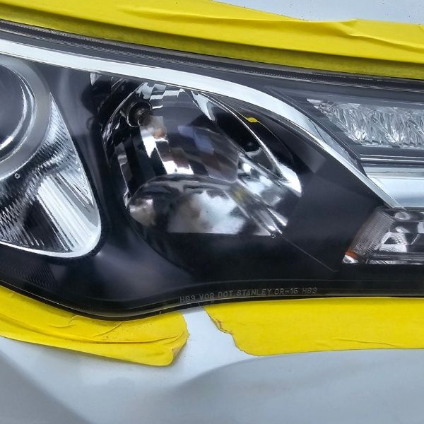A close-up of a car headlight housing, partially masked with yellow painter's tape for automotive repair.