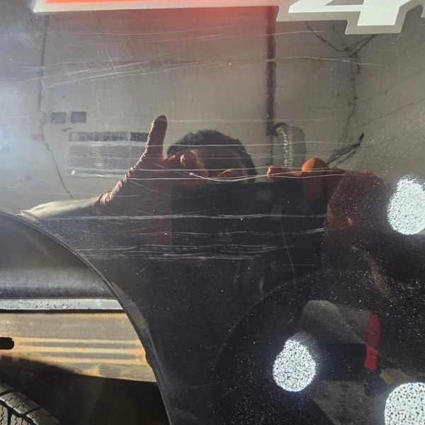 A reflection of a person taking a photo with their phone on a black, scratched surface, with part of a car emblem visible.