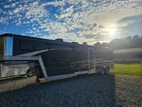 A black gooseneck trailer parked on a gravel lot under a bright, sunny sky with scattered clouds.