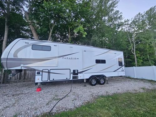 A white fifth-wheel travel trailer parked on a gravel lot next to a white fence and lush green trees.