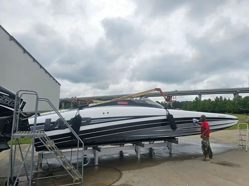 A person cleans the side of a sleek, black-and-white performance boat on a trailer parked outdoors near a bridge.