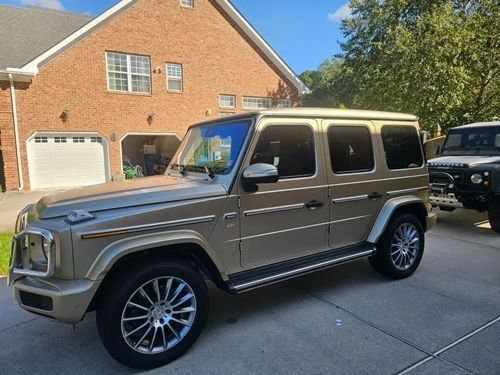 A gold Mercedes-Benz G-Class SUV parked on a residential driveway in front of a brick house.