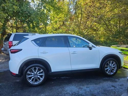 A white Mazda CX-5 SUV parked on a paved lot with trees in the background.