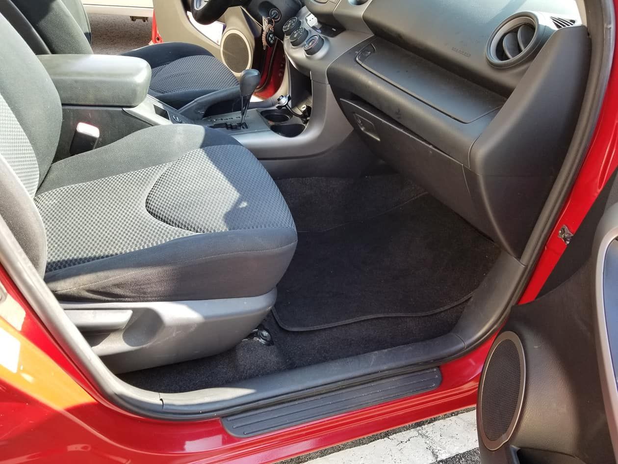 An interior view of a red vehicle showing the dark fabric passenger seat, dashboard, and carpeted floor mat.