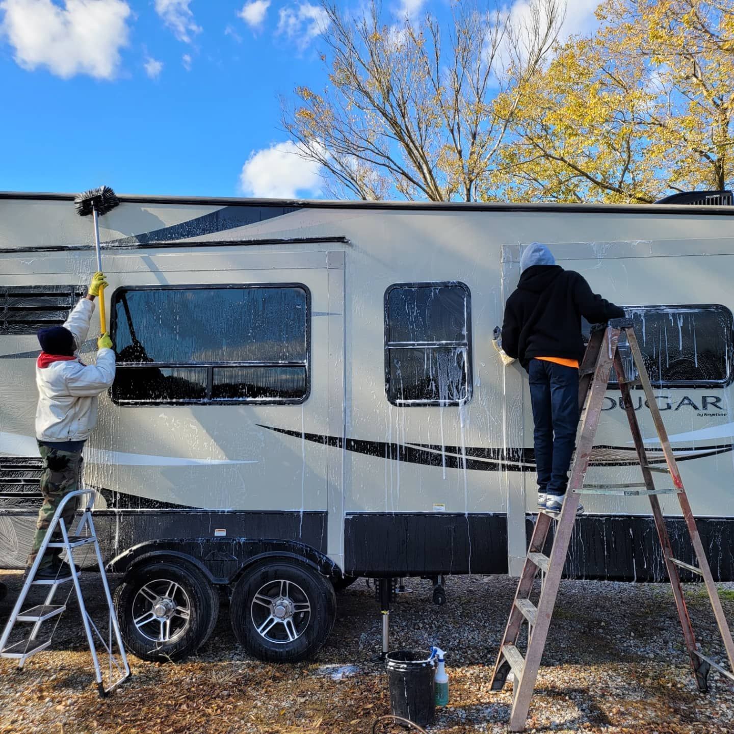 Two people on ladders wash the side of a large beige recreational vehicle with soap and brushes outdoors.