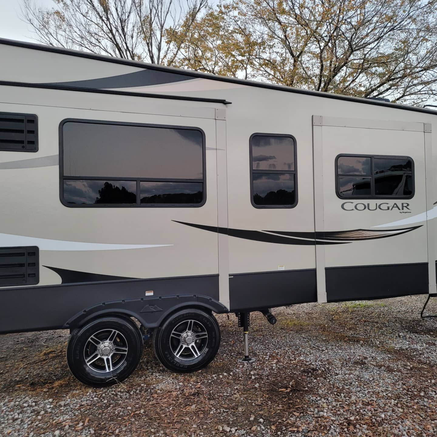 Tan and black Cougar travel trailer parked on a gravel surface outdoors with trees in the background.