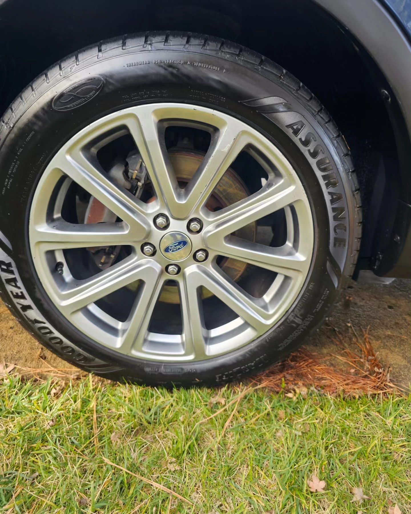 A close-up view of a silver, multi-spoke Ford alloy wheel with a Goodyear Assurance tire, parked on grass.