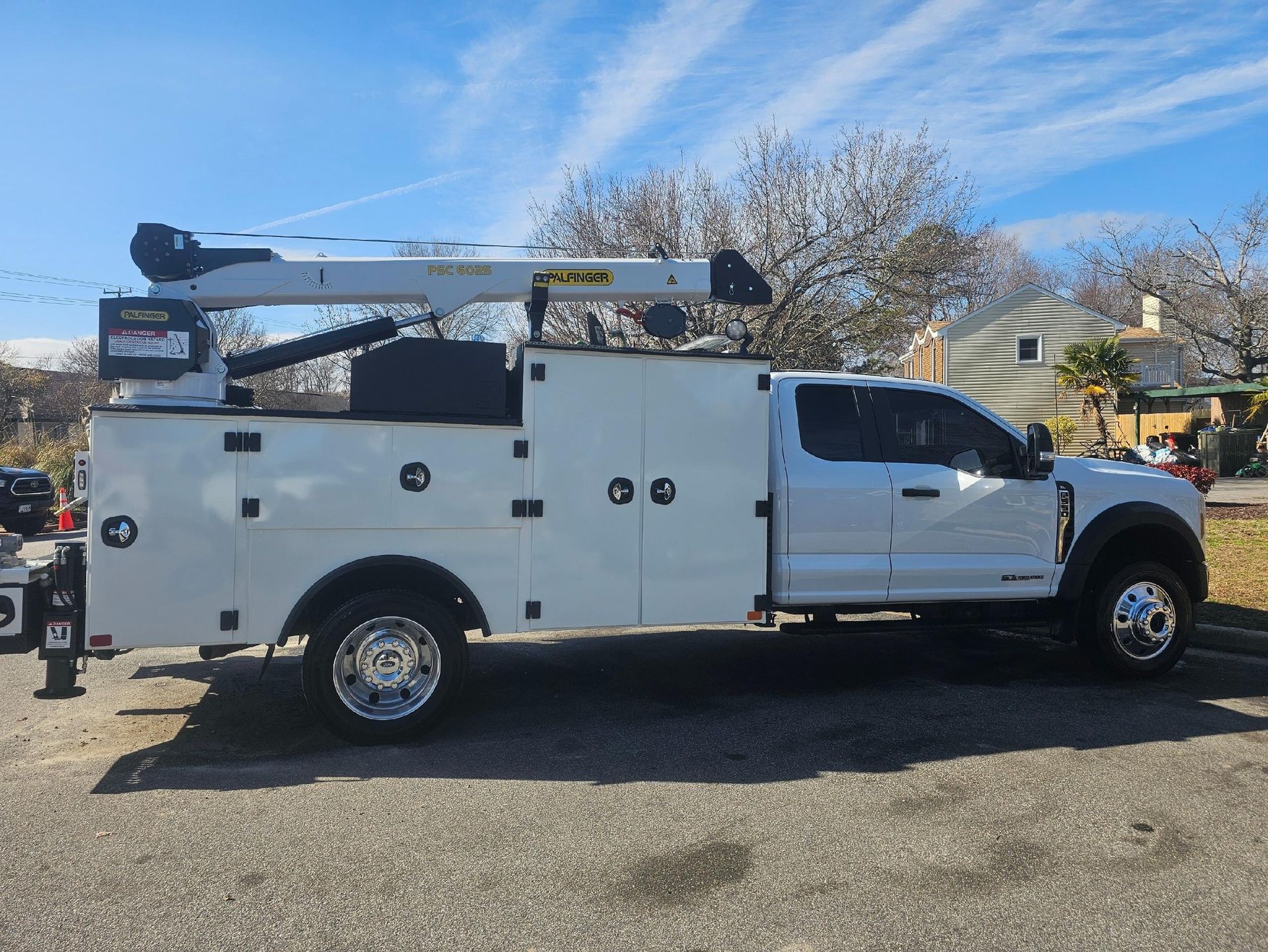 A white utility truck with a crane attachment parked on an asphalt lot against a sunny, blue sky.