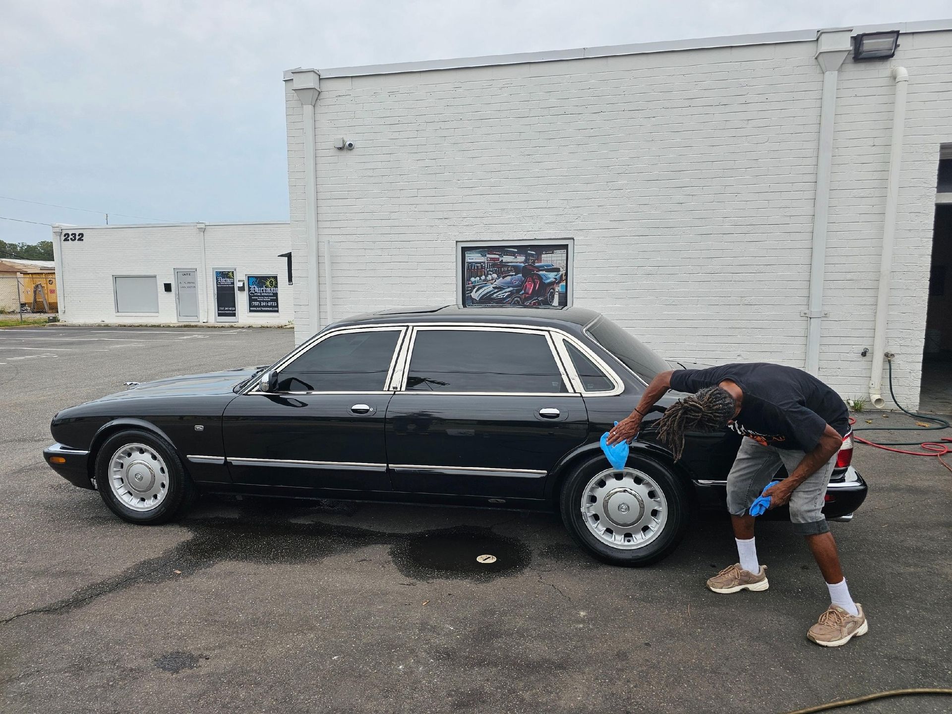 A person cleaning the rear wheel of a long, black stretch limousine parked in front of a white building.