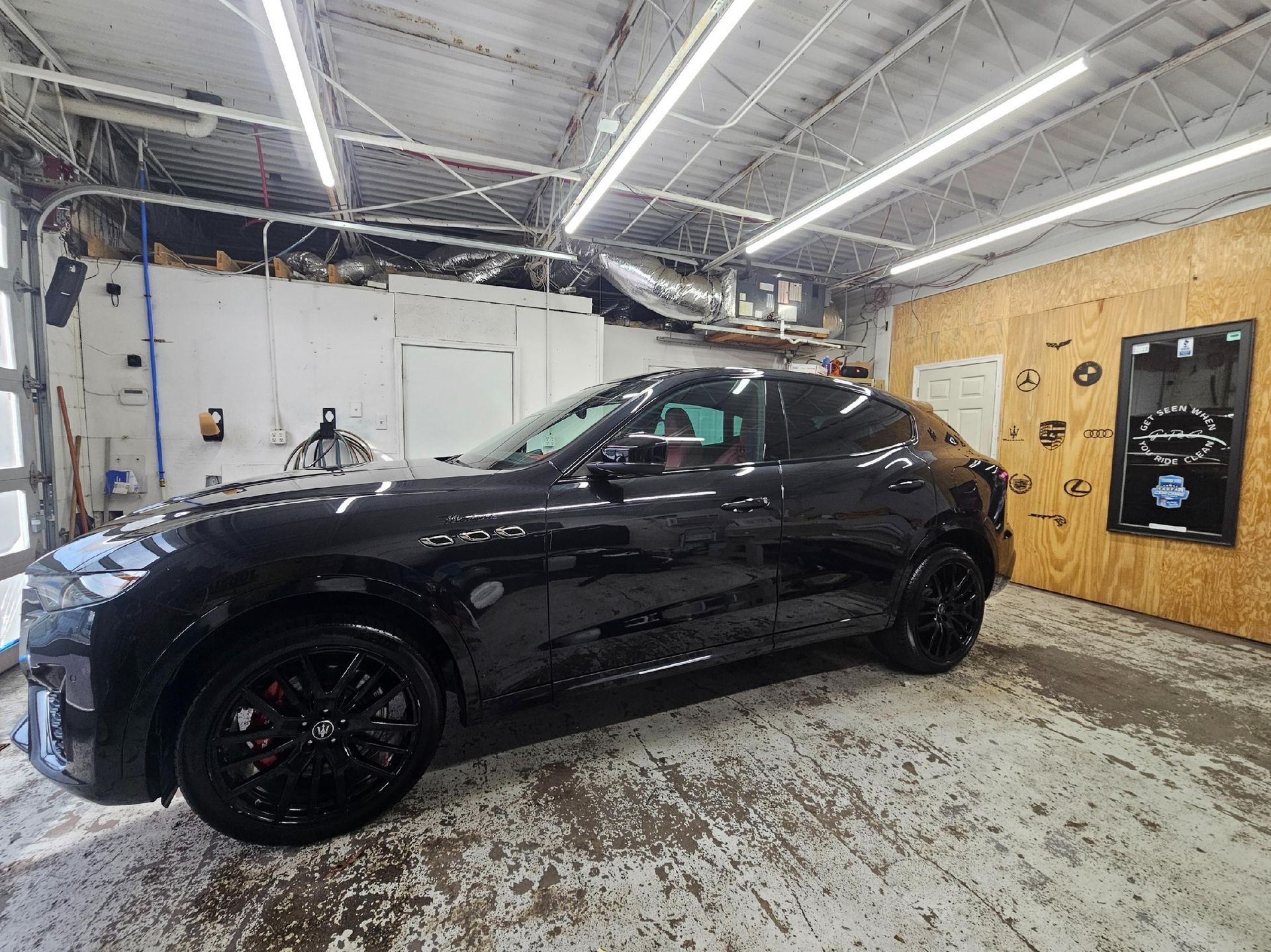 A glossy black SUV parked in a well-lit workshop with white walls and a plywood-covered wall with a framed sign.