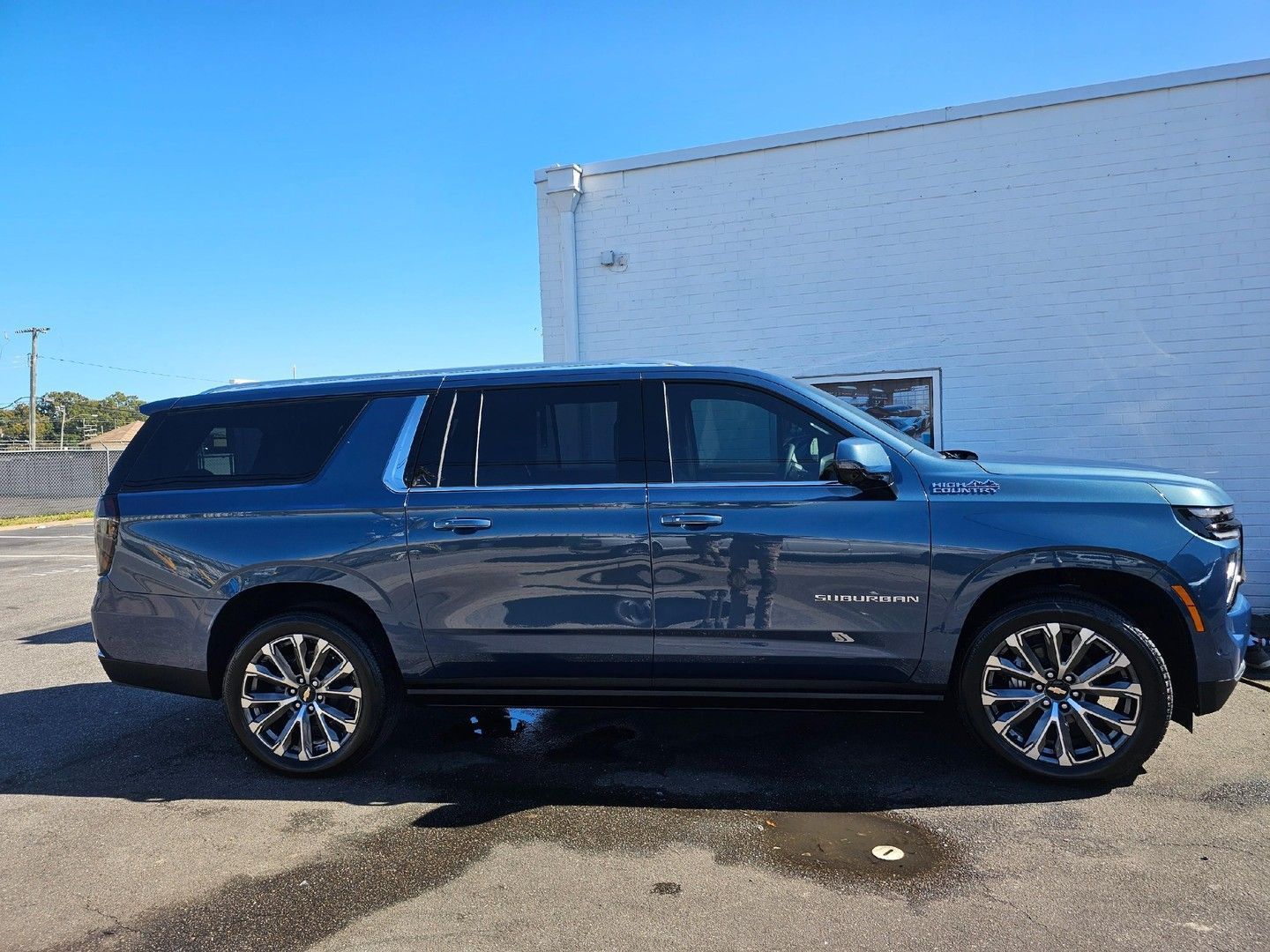 A blue Chevrolet Suburban parked on an asphalt lot beside a white building under a clear blue sky.
