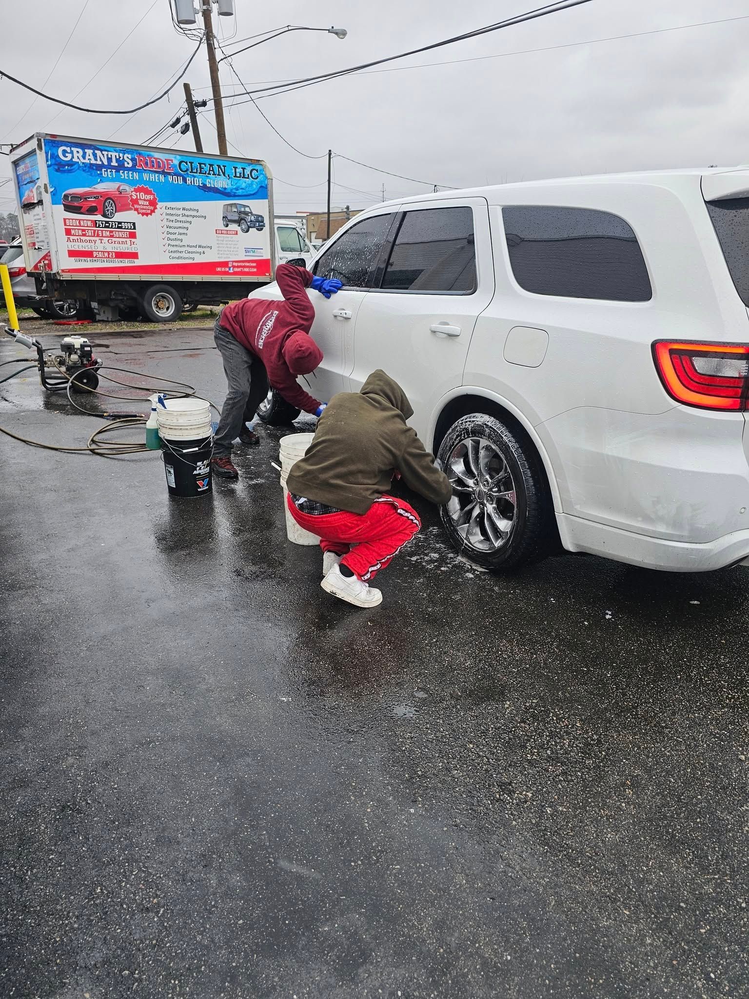 Two people wash a white SUV outdoors in a parking lot next to a mobile car wash service vehicle.