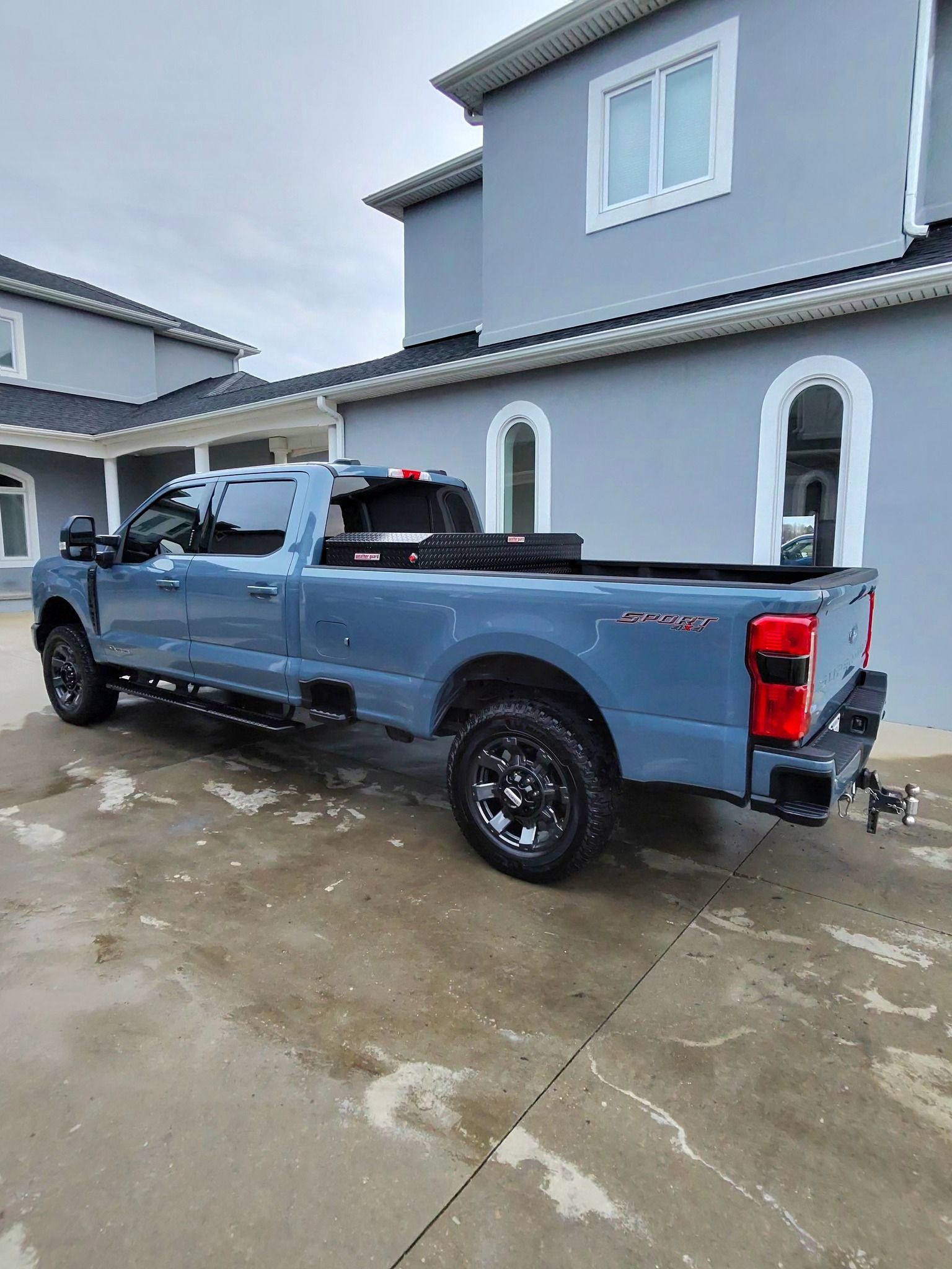 A light blue crew cab Ford Super Duty pickup truck with a black tool box in the bed, parked on a concrete driveway.