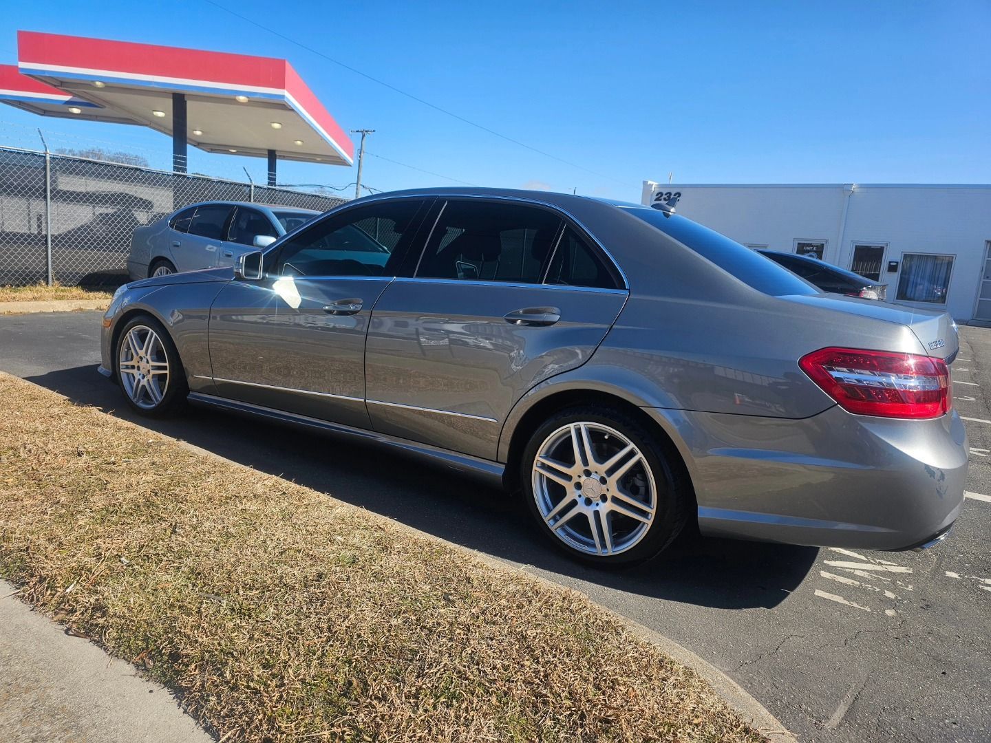 A grey Mercedes-Benz sedan parked on asphalt near a gas station on a sunny day.