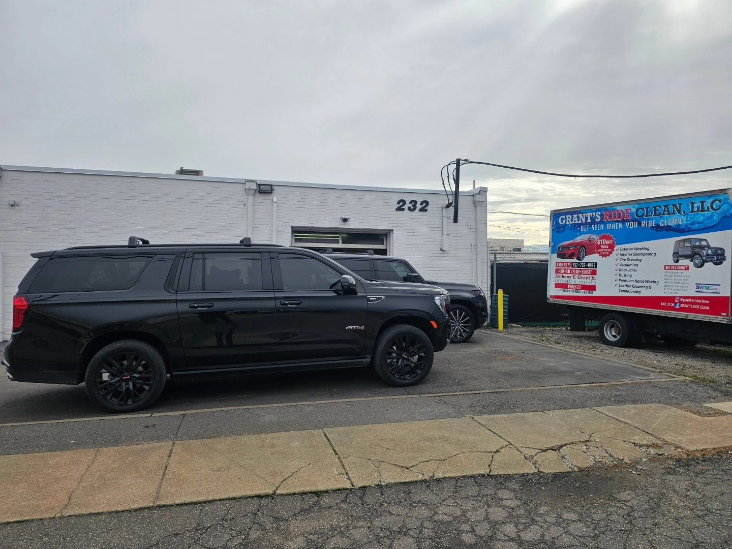 A black SUV parked in front of a white building numbered 232, with a branded box truck parked to the right.