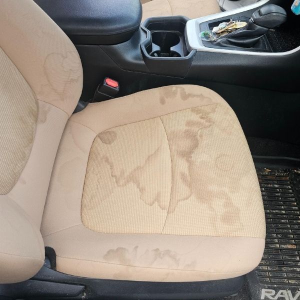 Light-colored fabric car seat stained with large, brownish liquid spills.