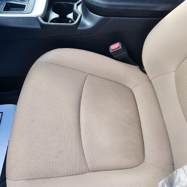 A tan fabric car seat with a seatbelt buckle visible, viewed from above next to the center console and cupholders.