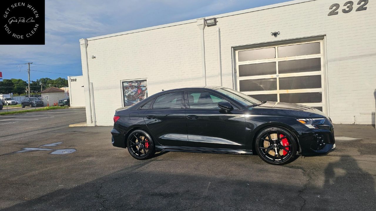 A black Audi sedan with red brake calipers parked outside a white industrial building on a sunny day.