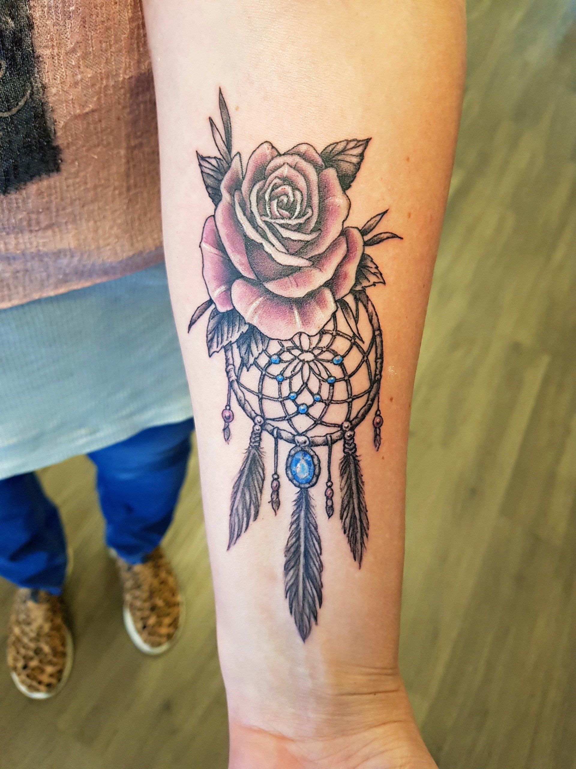 Close-up van een onderarm met een tatoeage van een roos en een dromenvanger in zwart en roze.