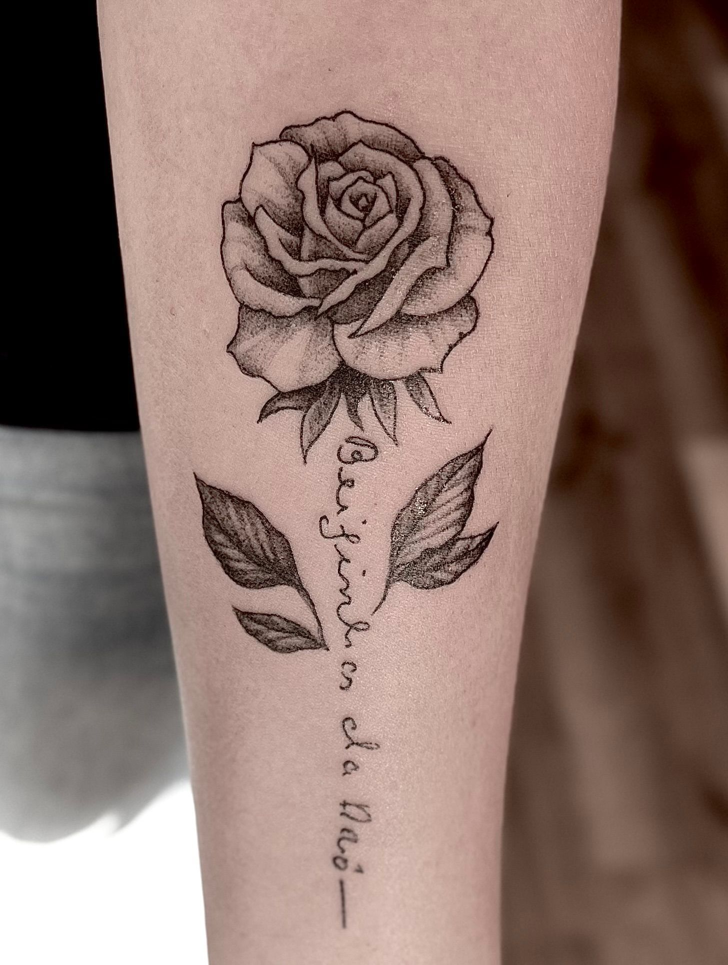 Zwart-grijze roos-tatoeage met bladeren en een klein citaat in sierletters op een onderarm.