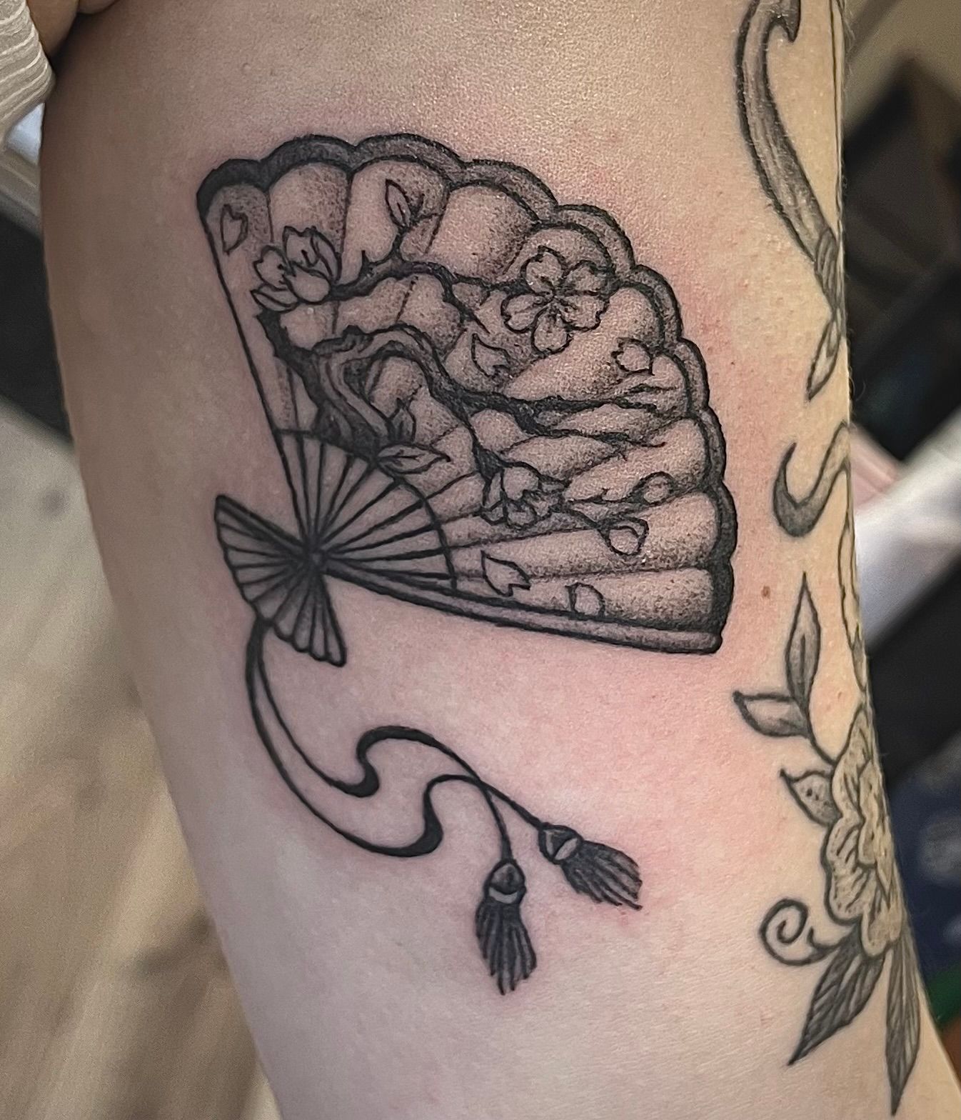 Zwarte contourtattoo van een handwaaier met bloemen op de huid, plus een gedeeltelijke bloementattoo in de buurt.