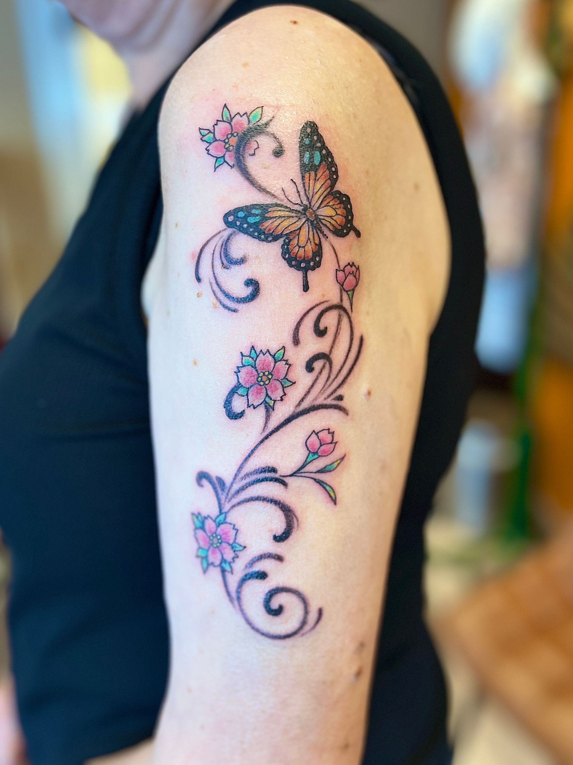 Tattoo op de bovenarm met een bloemenrank en een vlindermotief in roze, paars en oranje.
