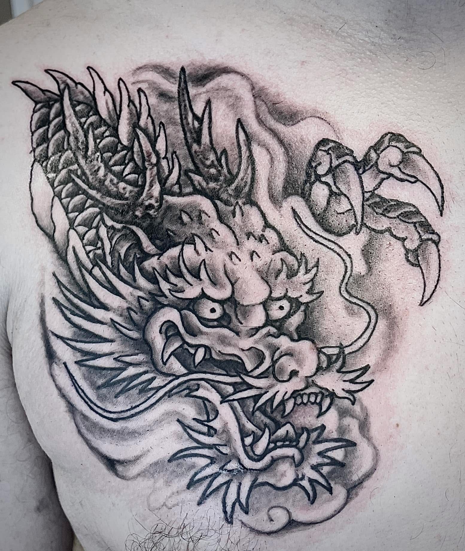 Zwart-grijze drakentattoo met open bek, gekrulde hoorns en een klauwpoot op de huid.