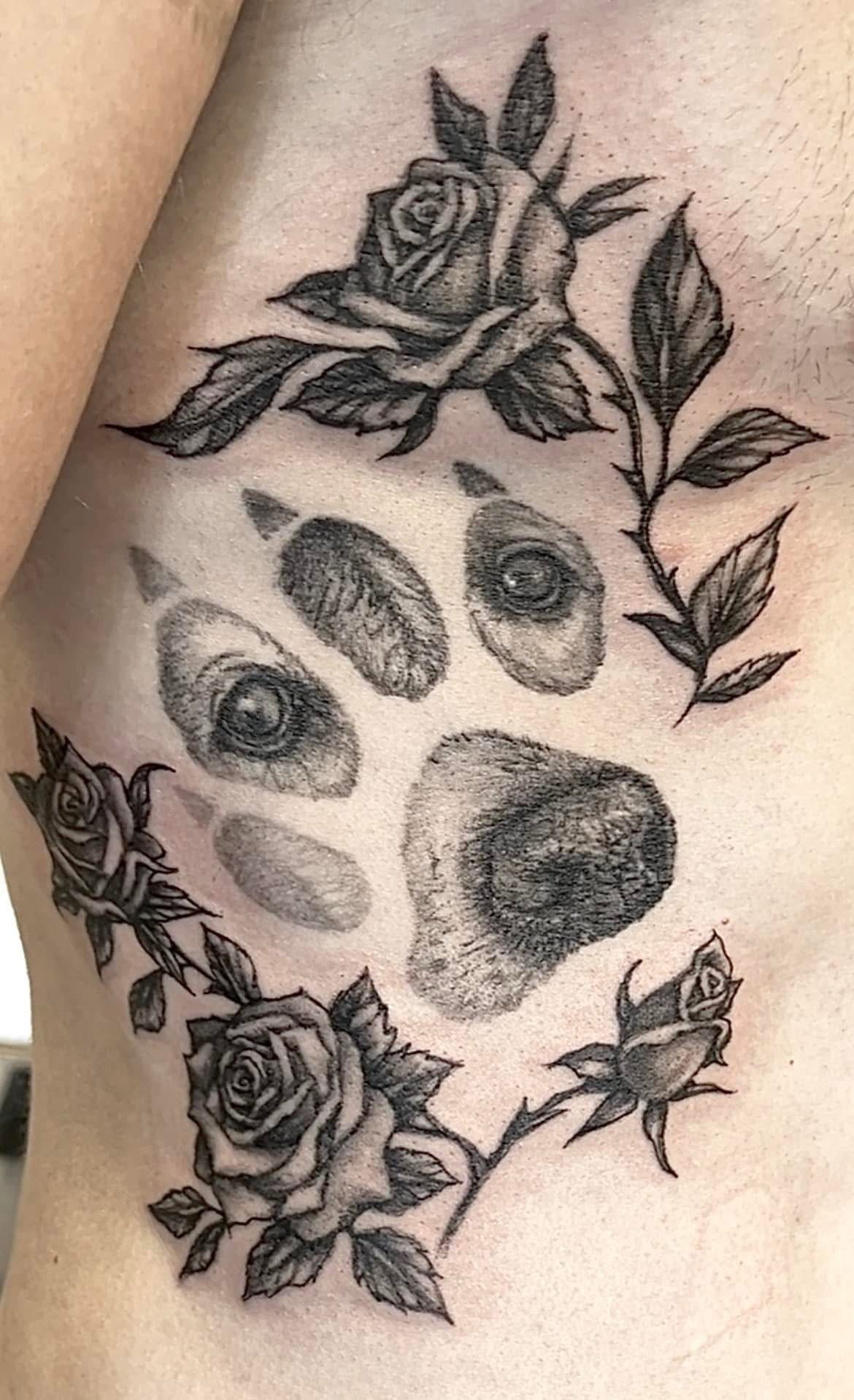 Zwart-grijze pootafdruktattoo omringd door rozen en bladeren op de huid