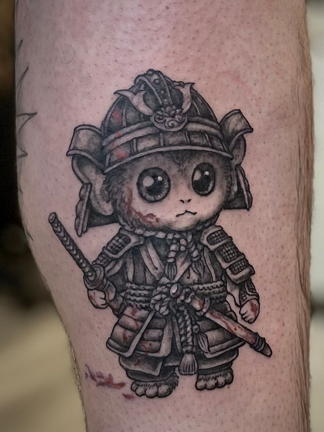 Schattige zwart-grijze tattoo van een chibi samurai-kat met helm, harnas en twee zwaarden op de huid.