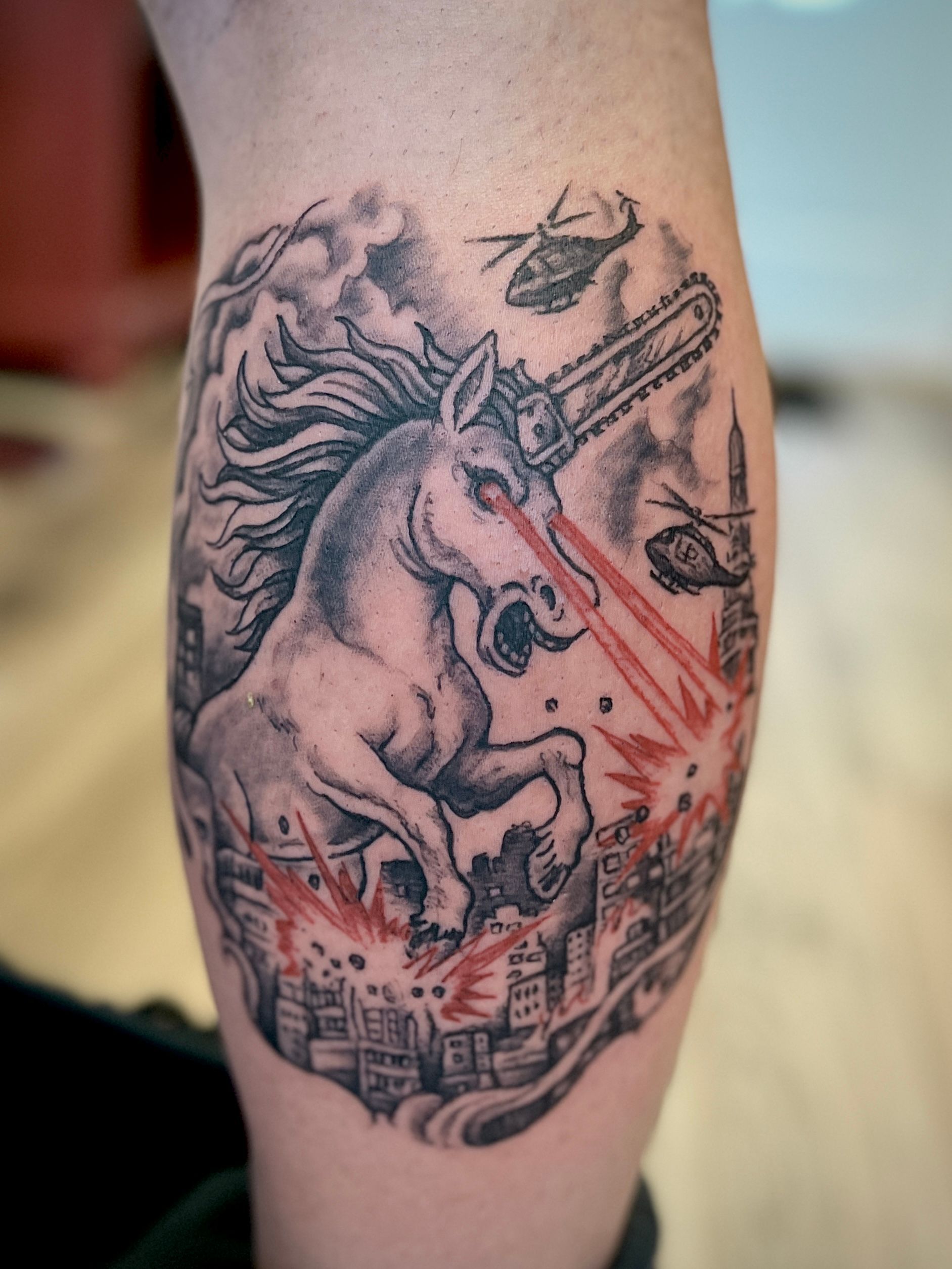 Armtattoo van een eenhoorn die een stadsgezicht doorboort, met rode accenten en een zwart-grijs ontwerp.