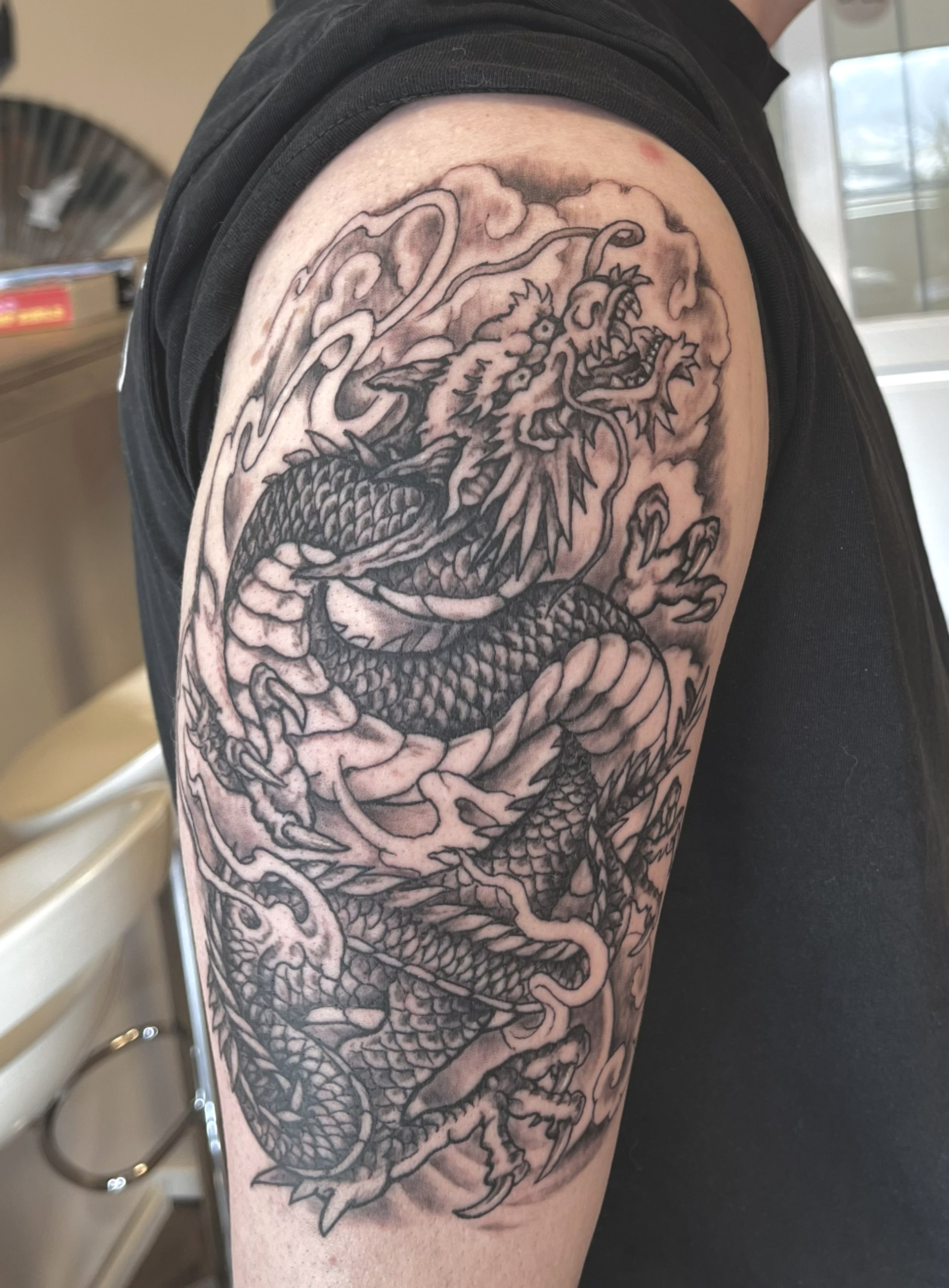 Zwart-grijze drakentattoo die de bovenarm bedekt, met wervelende wolkdetails en gedurfde lijnen.