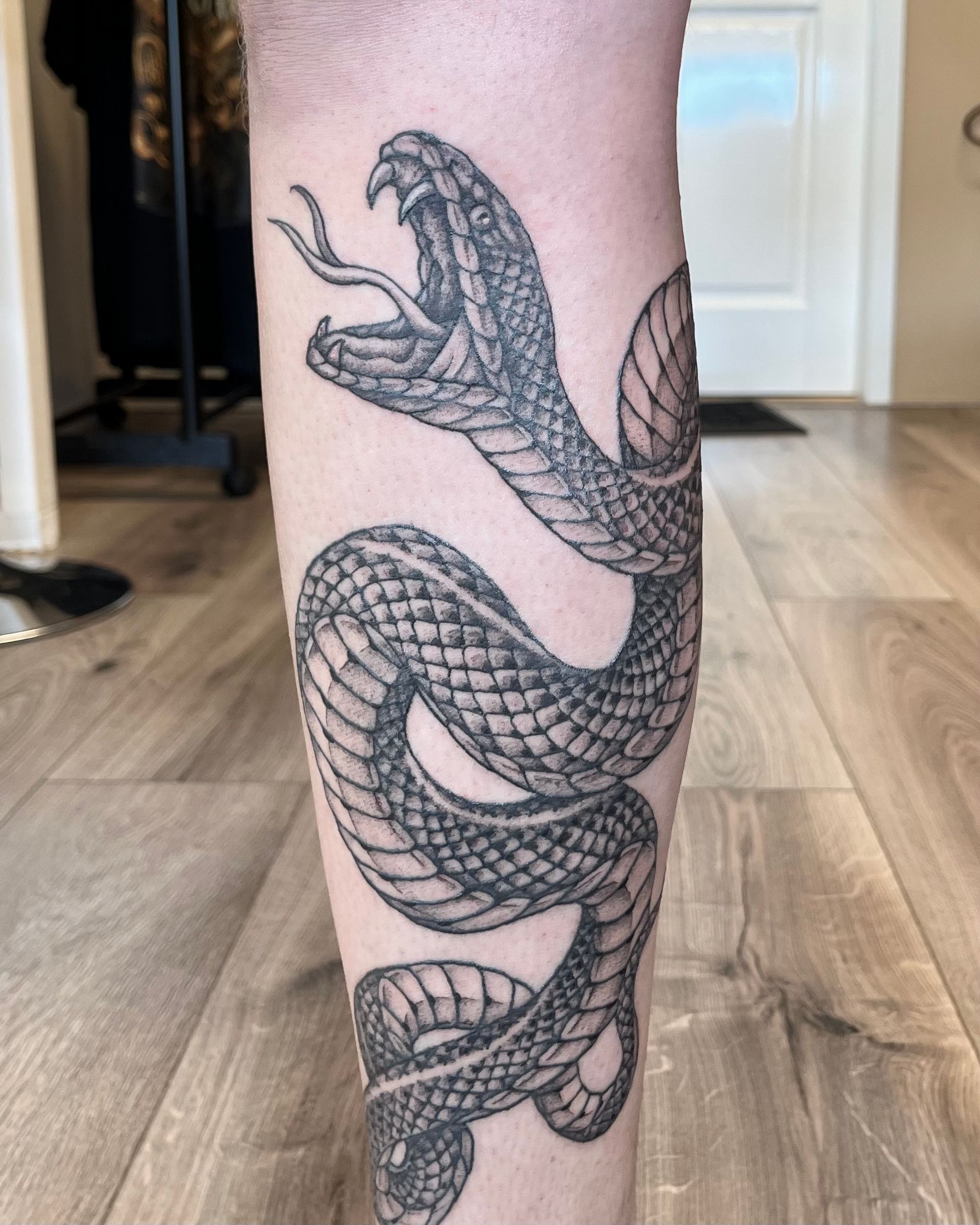 Een zwarte slangentattoo die zich om het onderbeen wikkelt, met gedetailleerde schubben en een open bek.